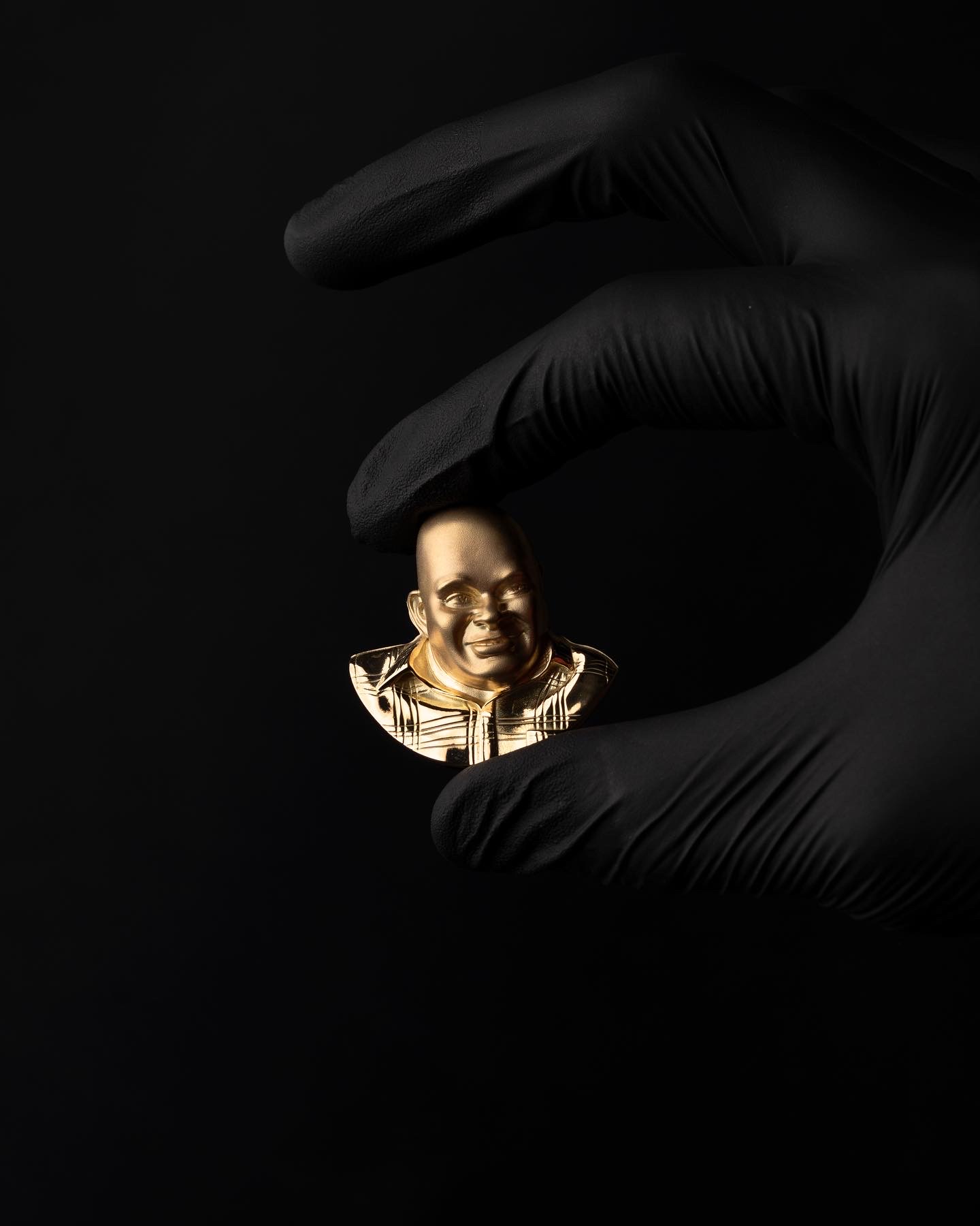 Finished gold three-dimensional portrait pendant held between black-gloved fingers